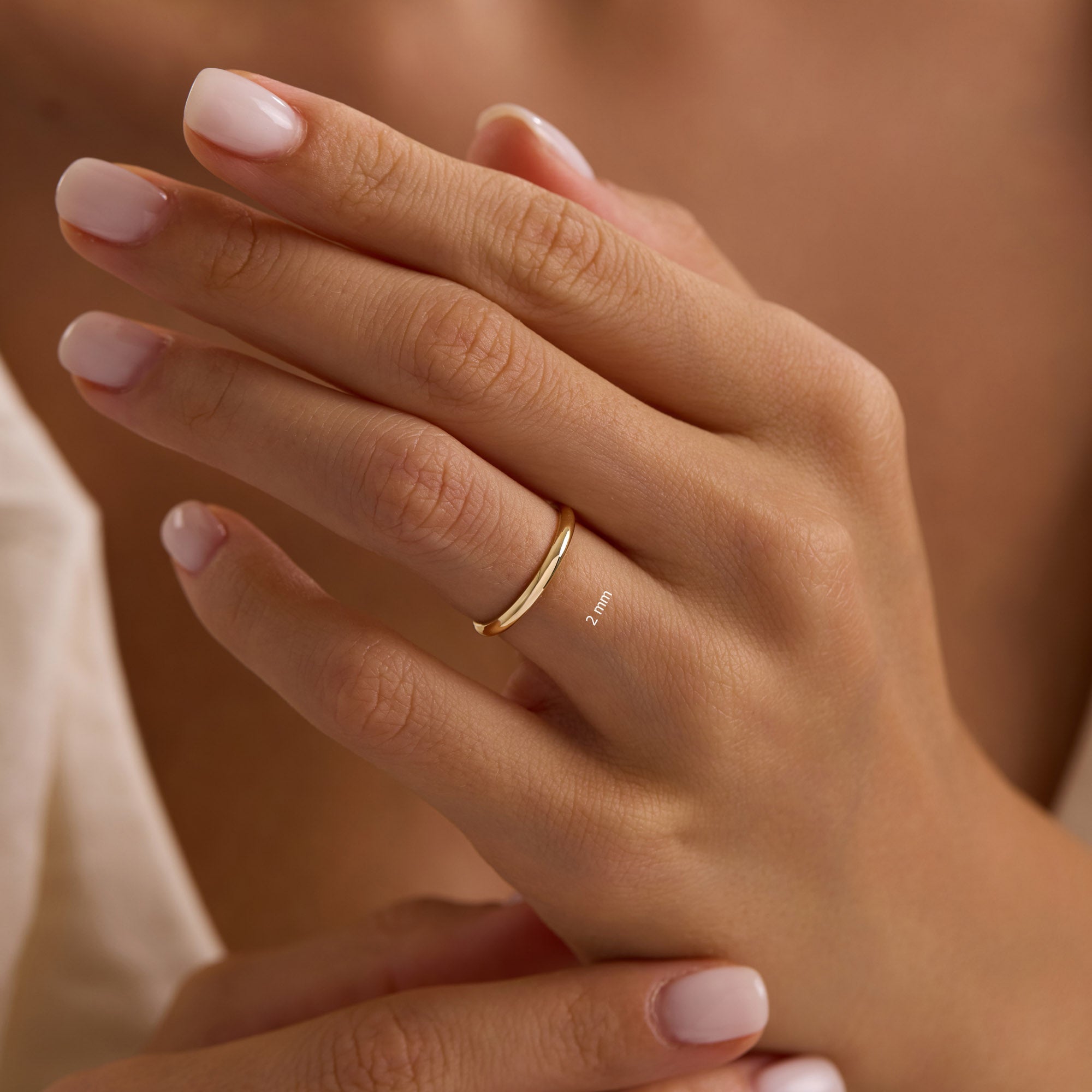 A close-up of a hand wearing a thin 2 mm gold band on the ring finger. The nails are neatly manicured with a soft pink polish, and the background is softly blurred, highlighting the delicate ring.