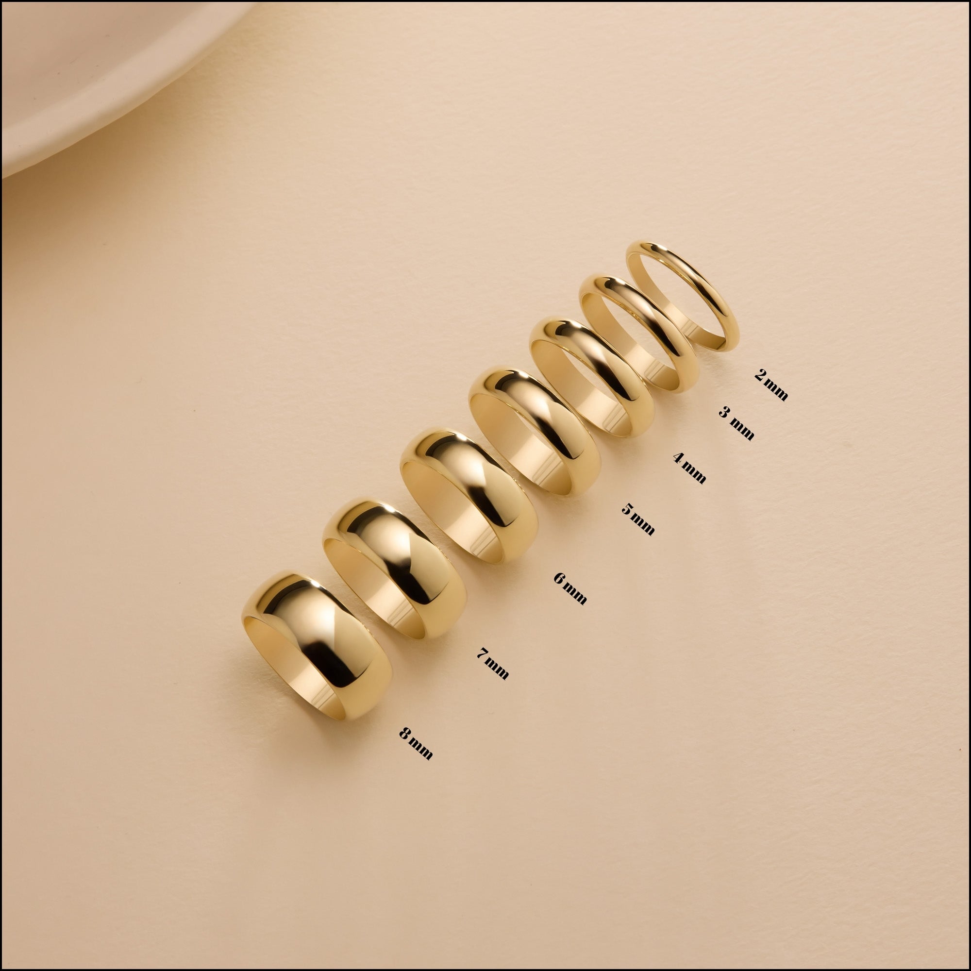 A row of gold dome-shaped rings arranged from widest to thinnest on a beige surface. Each ring is labeled with its band width, ranging from 8 mm down to 2 mm, creating a visual comparison of the different sizes.