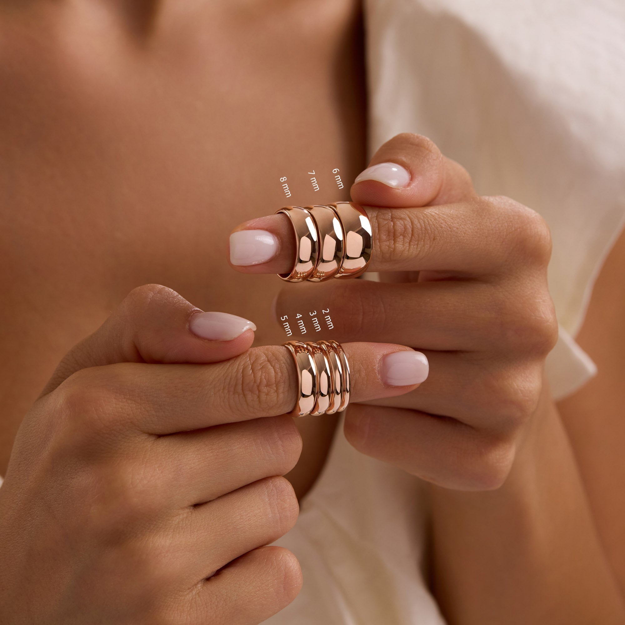 Model holding 14k solid rose gold wedding bands in multiple widths stacked on both thumbs, showing 2mm to 8mm dome bands.